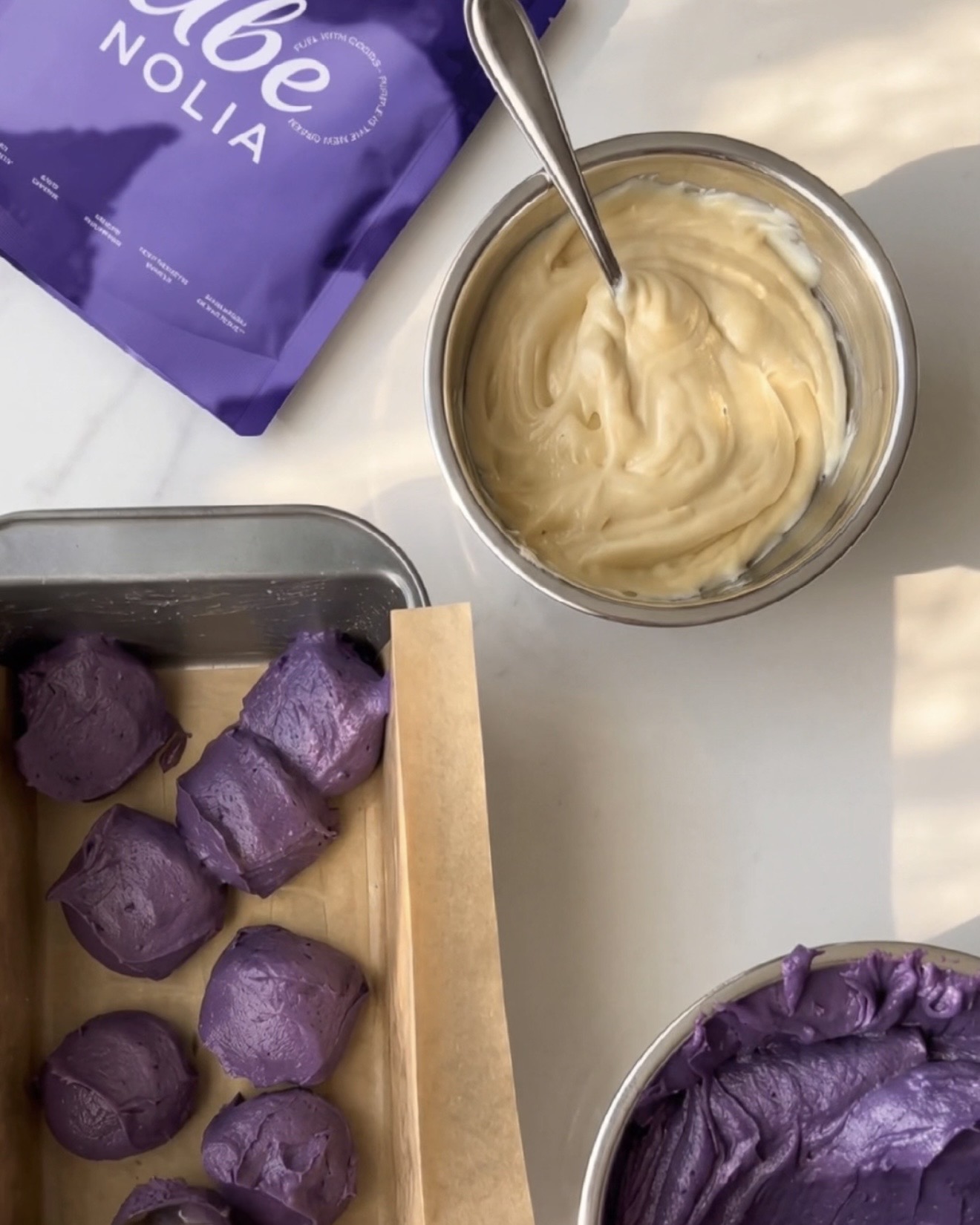Færdig hjemmelavet ube halaya, vaniljedej og skefulde lilla ube-dej i en brødform ved siden af en pose Ube Nolia ubepulver